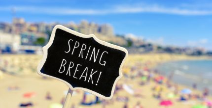 closeup of a black signboard with the text spring break written in it, in front of a blurred beach with many people bathing and sunbathing